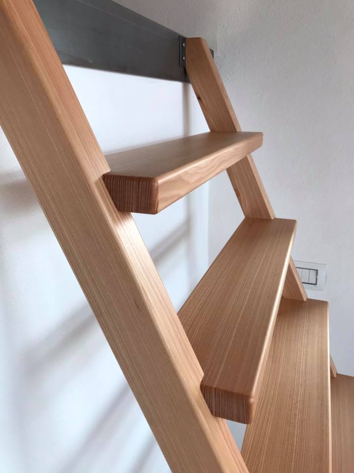 Primo piano di una scala in legno con ringhiere e gradini inclinati, sullo sfondo di una parete bianca.