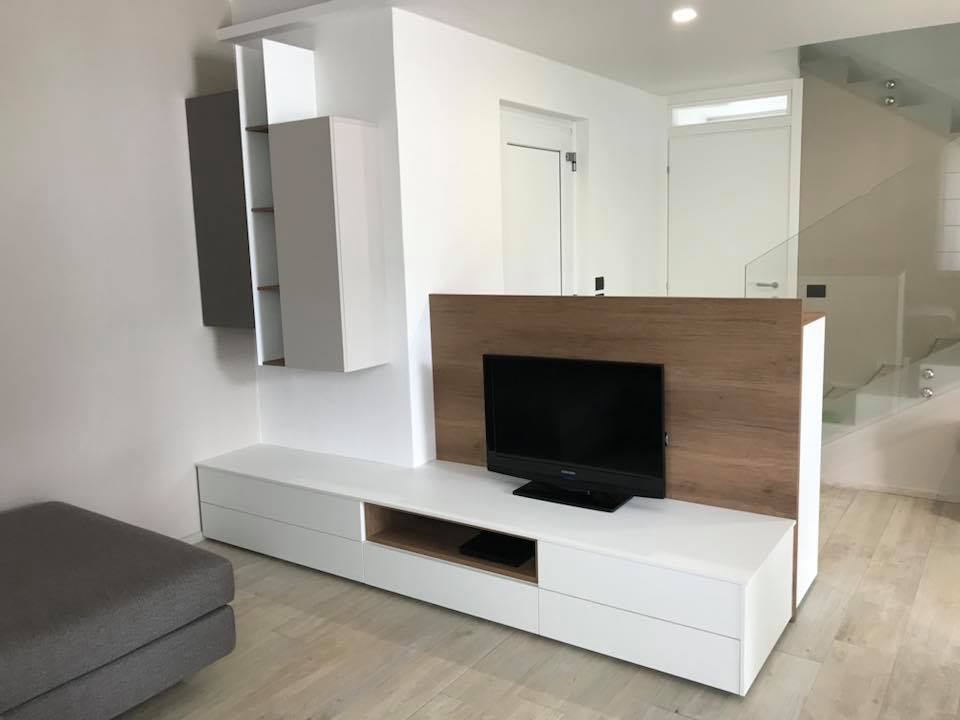 Soggiorno moderno con mobile TV bianco, pannello in legno, divano grigio e scala.