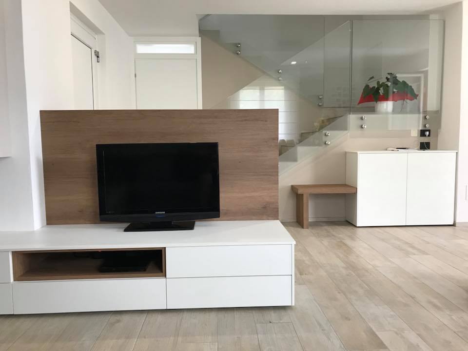 Soggiorno moderno con mobile TV bianco, pannello in legno, TV a parete e scale sullo sfondo.