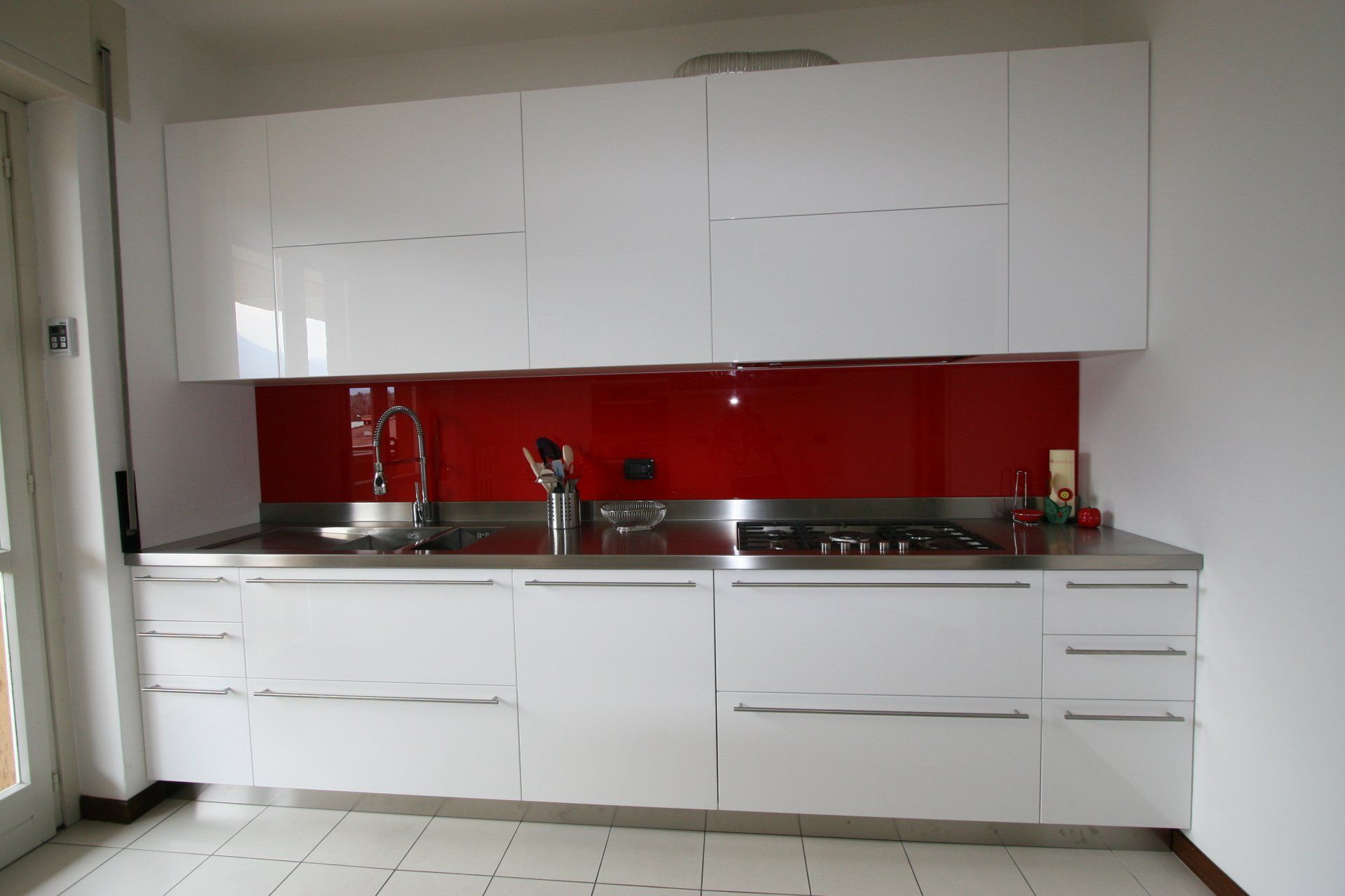 Cucina moderna bianca con paraschizzi rosso, lavello in acciaio inox e forno a incasso.
