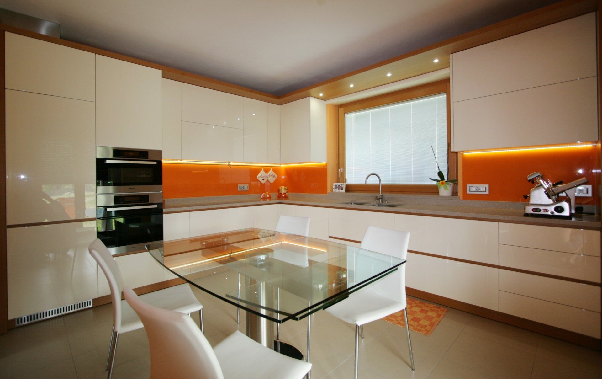 Cucina moderna con mobili bianchi, paraschizzi arancione, tavolo da pranzo in vetro e grande finestra sopra il lavello.