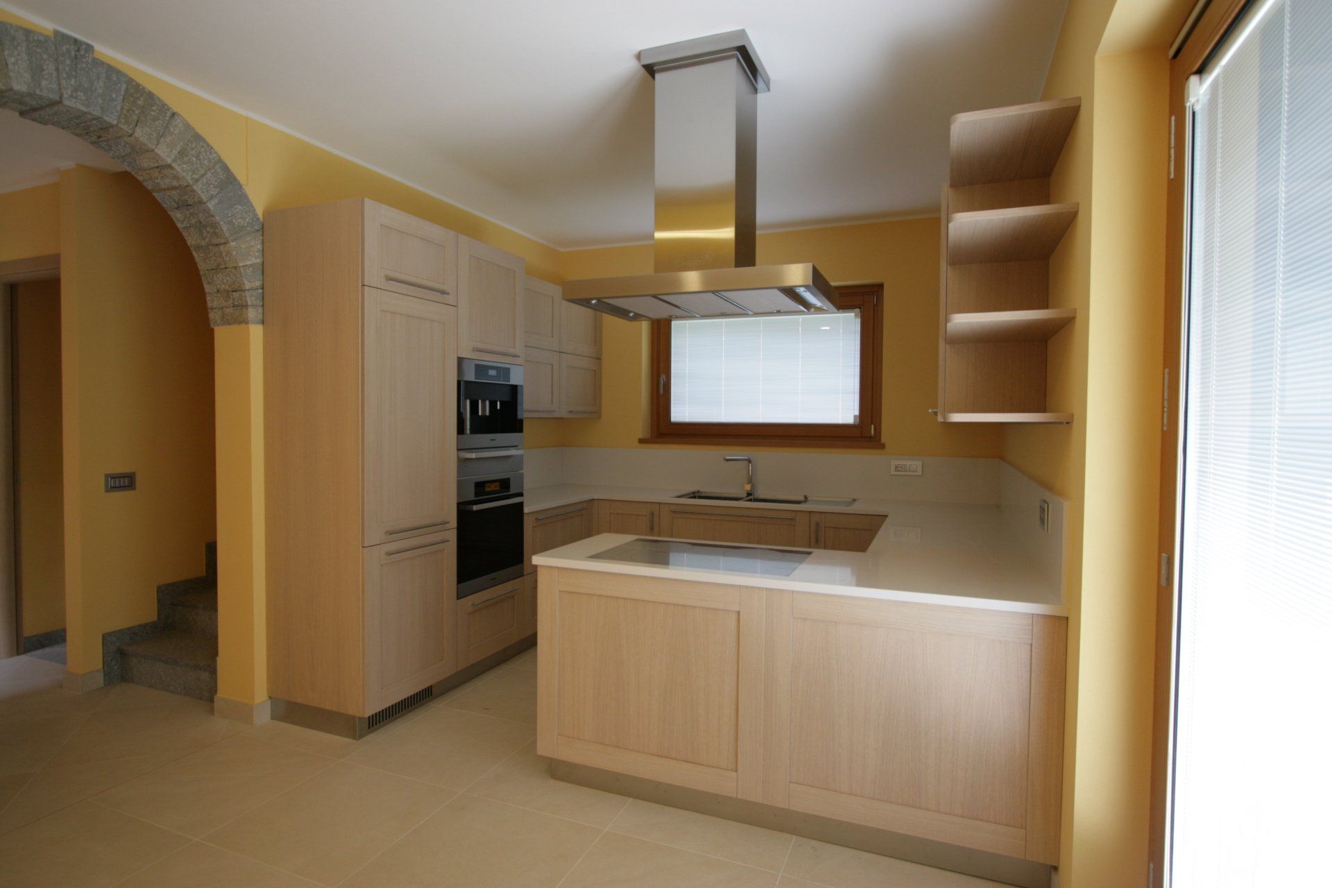 Cucina luminosa con mobili in legno chiaro, isola centrale, cappa in acciaio inox, porta ad arco e finestra soleggiata.