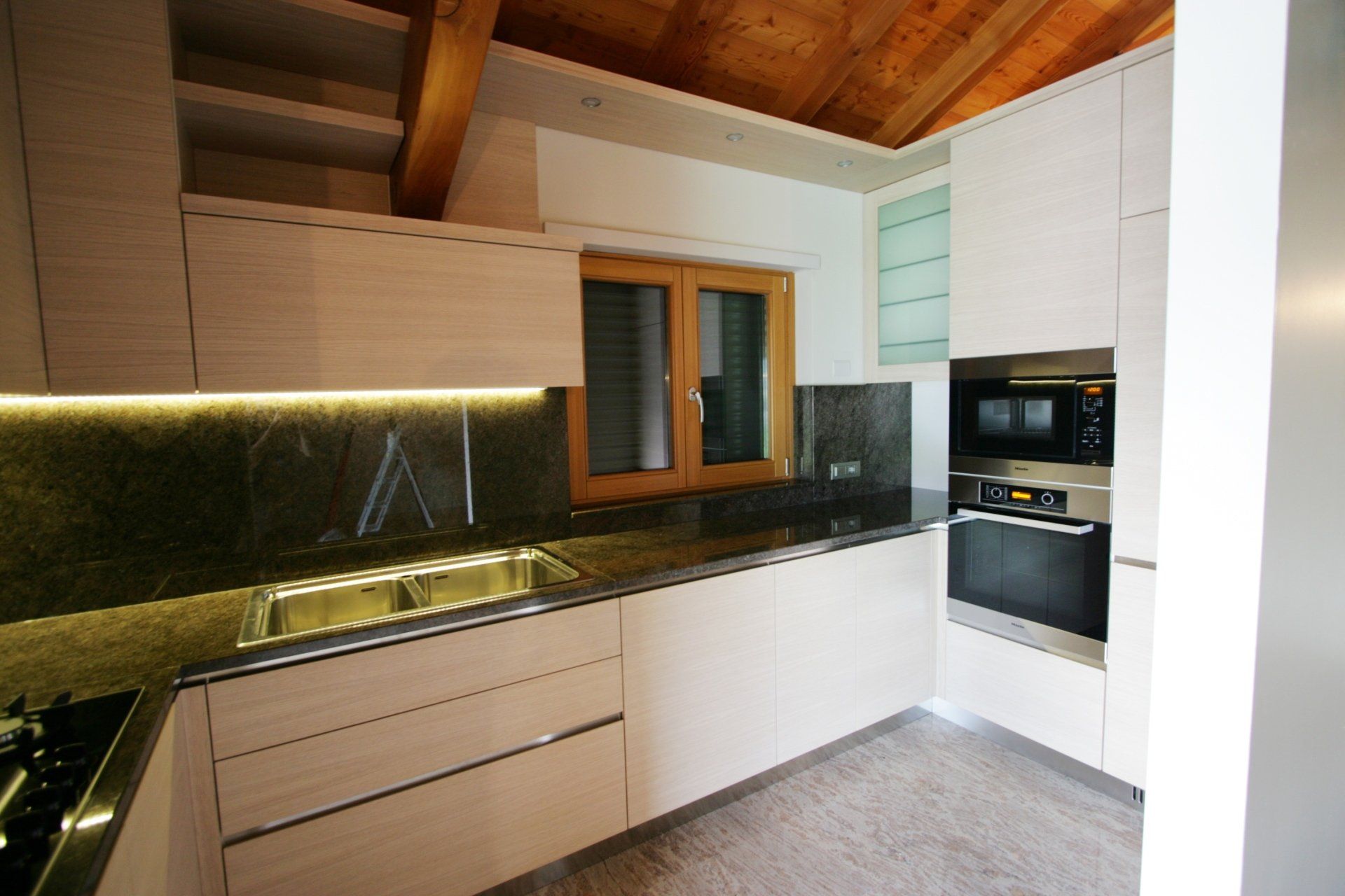 Cucina moderna con mobili bianchi, ripiani scuri, soffitto in legno ed elettrodomestici a incasso.
