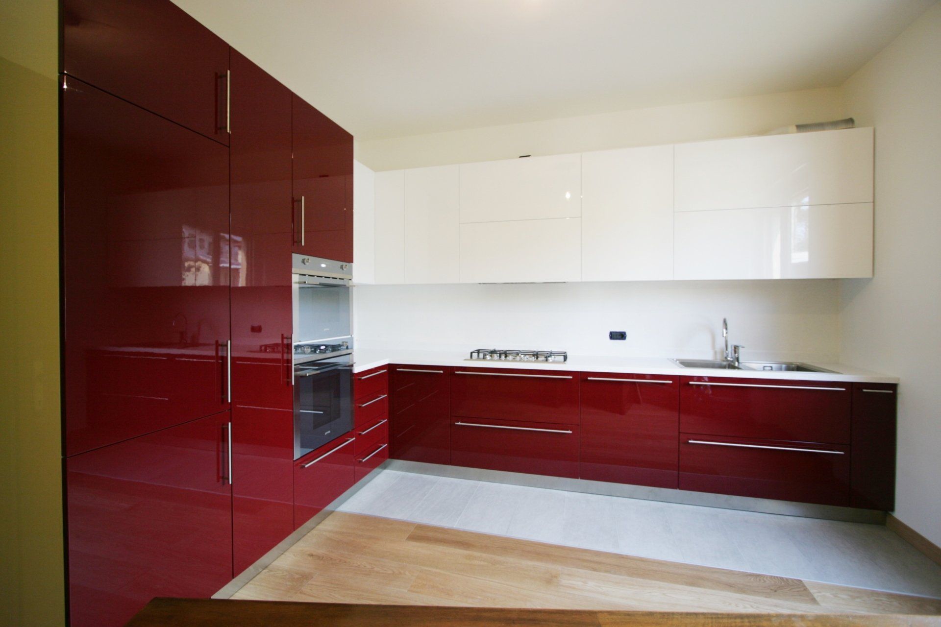 Cucina moderna con mobili lucidi rossi e bianchi, forno a incasso e pavimento in legno chiaro.
