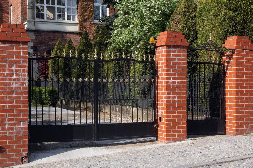 A wrought iron gate is surrounded by brick pillars in front of a brick house.