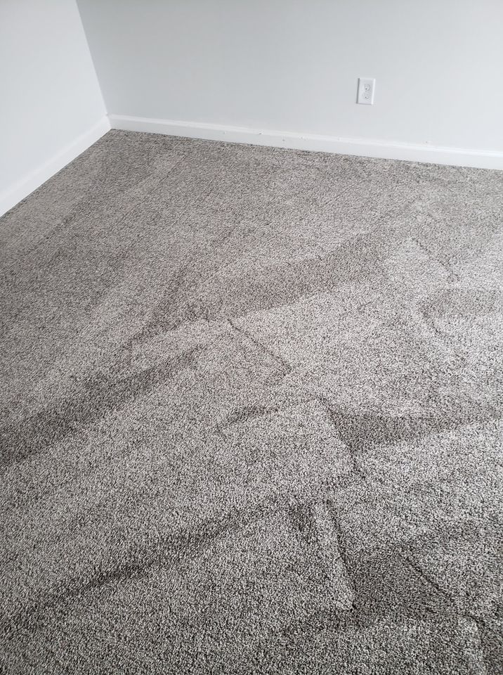 Gray carpet in a corner of a room, with ripples and indentations. White walls and an electrical outlet are visible.