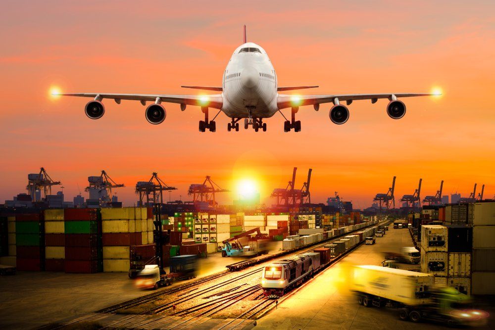 Cargo plane landing over a busy port with containers, trains, trucks, and a sunset.