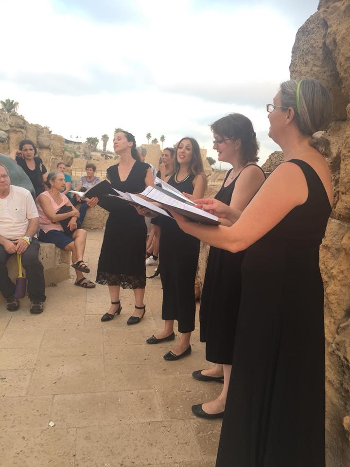 מקהלה בשמלות שחורות שרה בחוץ במרפסת אבן ציורית, כשקהל יושב בקרבת מקום