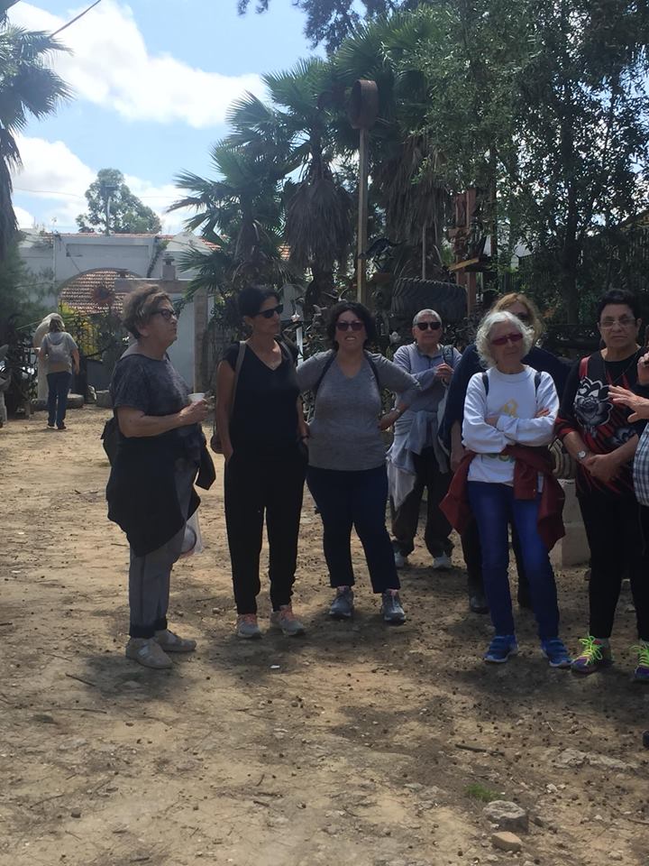 קבוצת אנשים עומדים על שביל עפר בחוץ ליד עצים ומבנים, משוחחים באור השמש.