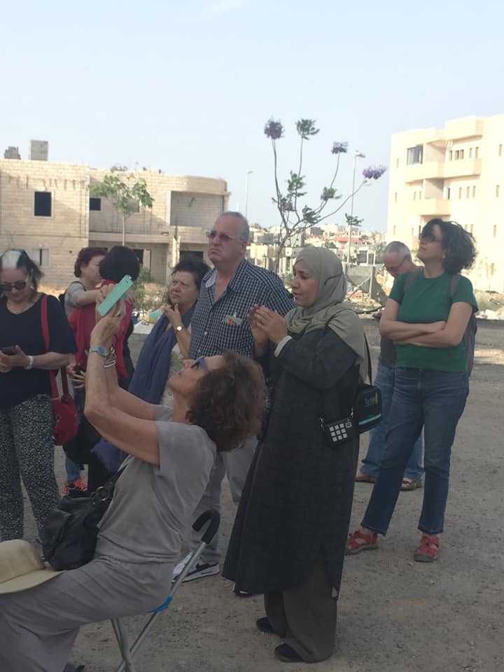 קבוצת אנשים בחוץ בשקיעה, אחד יושב ומצלם בעוד אחרים עומדים ומשוחחים ליד בנייני דירות.