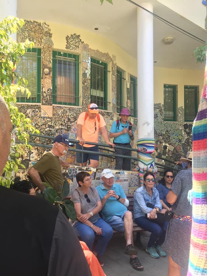 אנשים יושבים ועומדים מחוץ לבניין עם תריסים ירוקים, בחצר מוצלת.