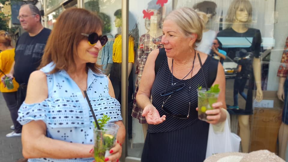 שתי נשים משוחחות בחוץ, כל אחת מחזיקה משקה ירוק, מול חלון ראווה של חנות.