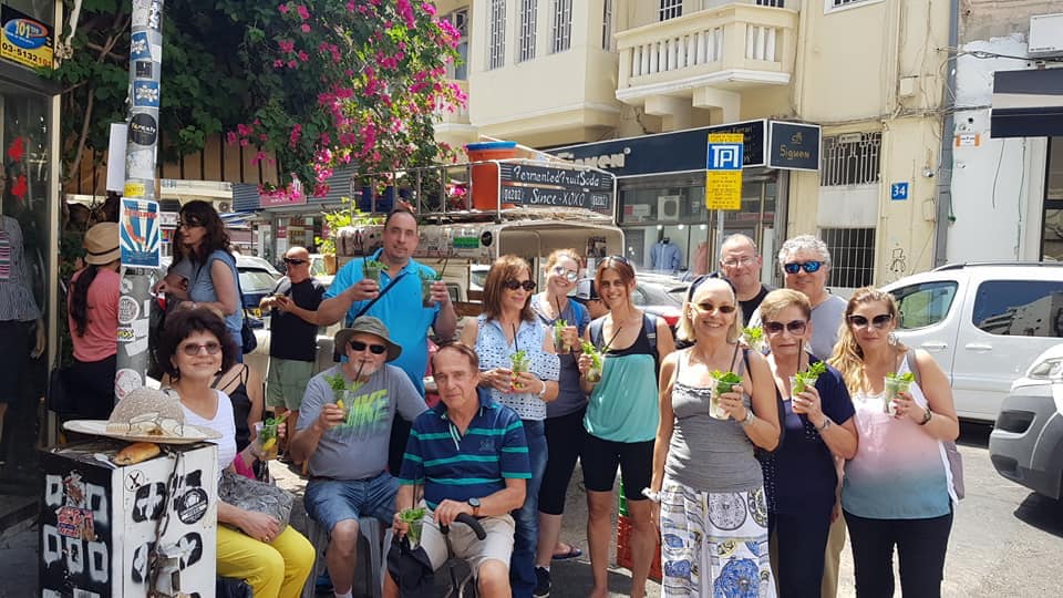 קבוצת תיירים הולכת ברחוב שטוף שמש, כמה מהם מחזיקים משקאות, כשחנויות ופרחים פורחים בקרבת מקום