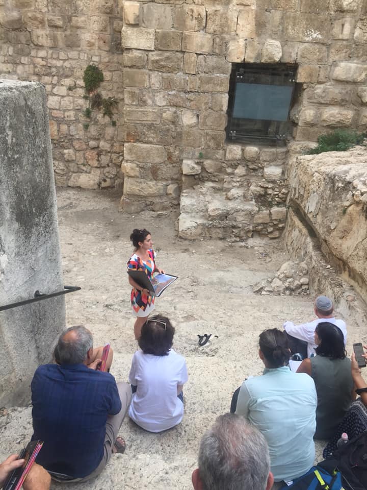 ילד קטן מדבר לקהל יושב בחצר אבן, אוחז בספר או נייר צבעוני.