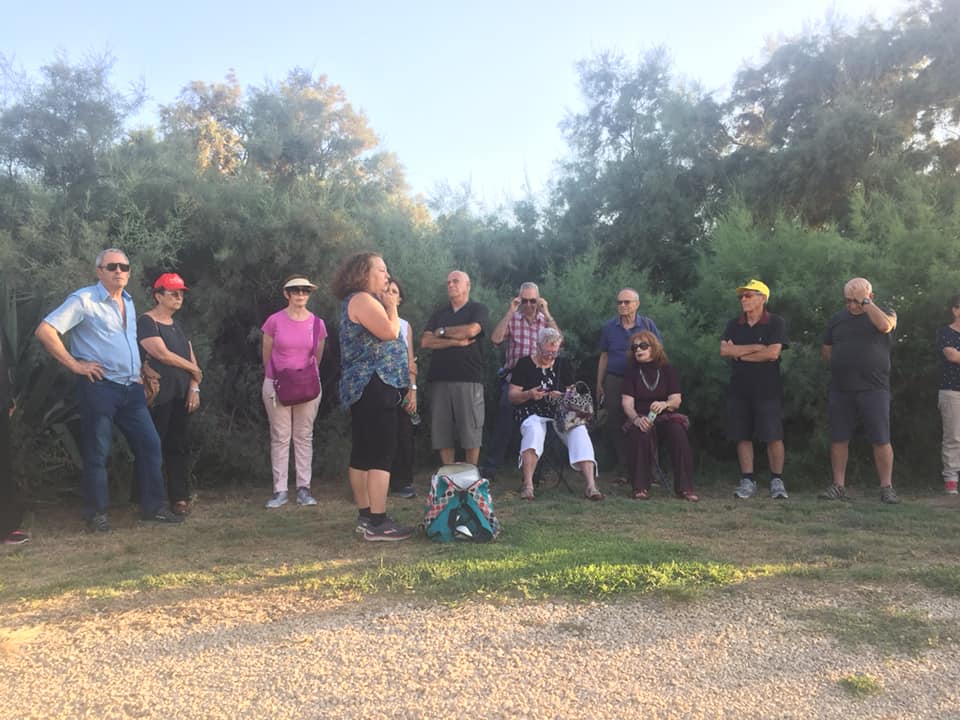 קבוצת אנשים עומדת בחוץ על שביל עשבוני עם עצים ברקע, חלקם חובשים כובעים ומשקפי שמש