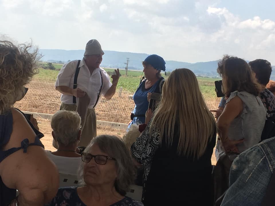 קבוצה מאזינה לרמקול בחוץ מתחת לאוהל, כשברקע נראות גבעות.