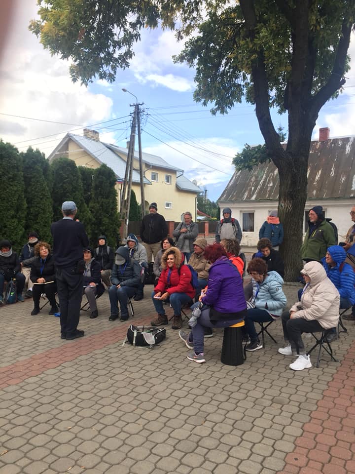 אנשים יושבים בחוץ בכיכר, מאזינים לרמקול ליד עץ ביום מעונן.
