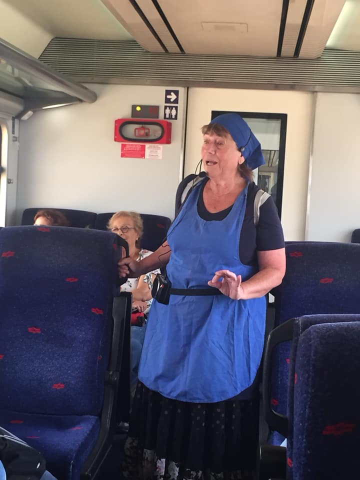 אישה בסינר כחול ומצנפת עומדת ומדברת במעבר רכבת בין המושבים