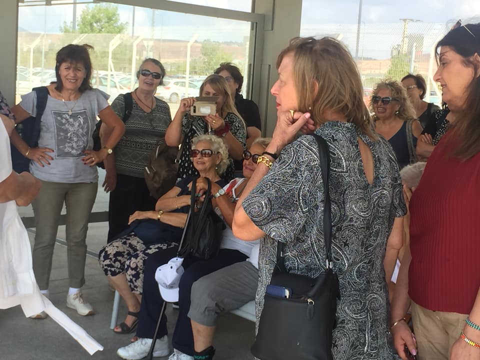 קבוצת אנשים מתרועעים בתוך הבית, כאשר אישה אחת יושבת ואחרות עומדות סביבה בחדר מואר.