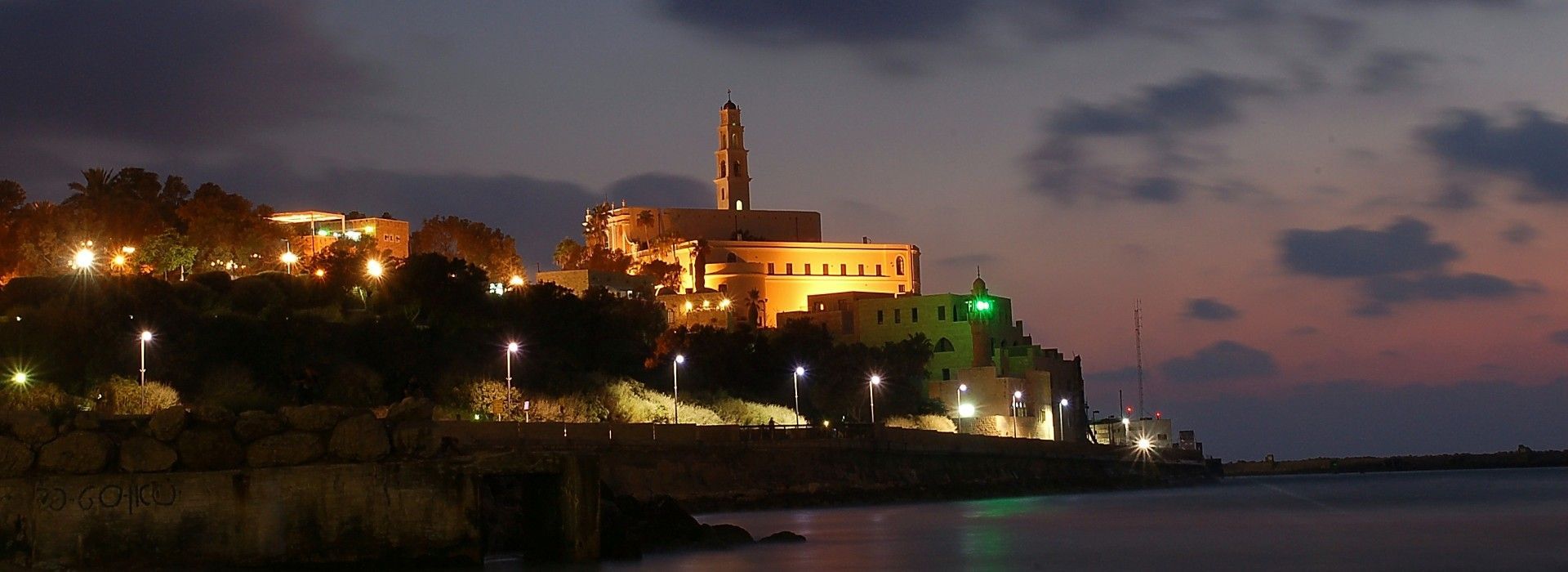עיירת חוף עם רדת החשיכה עם בניינים זוהרים, מגדל מואר ומים שקטים תחת שמיים סגולים