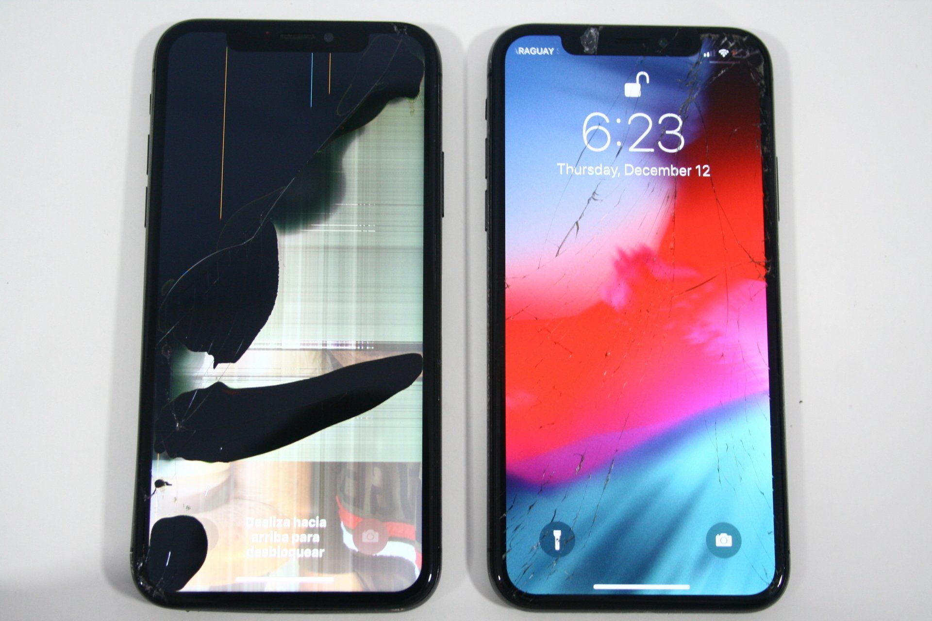 Two cell phones are sitting next to each other on a table with broken screens. The one on the left looks much more broken.