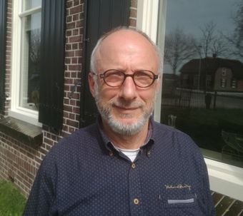 Een persoon met een bril en een baard staat lachend buiten voor een bakstenen gebouw met een groot raam.