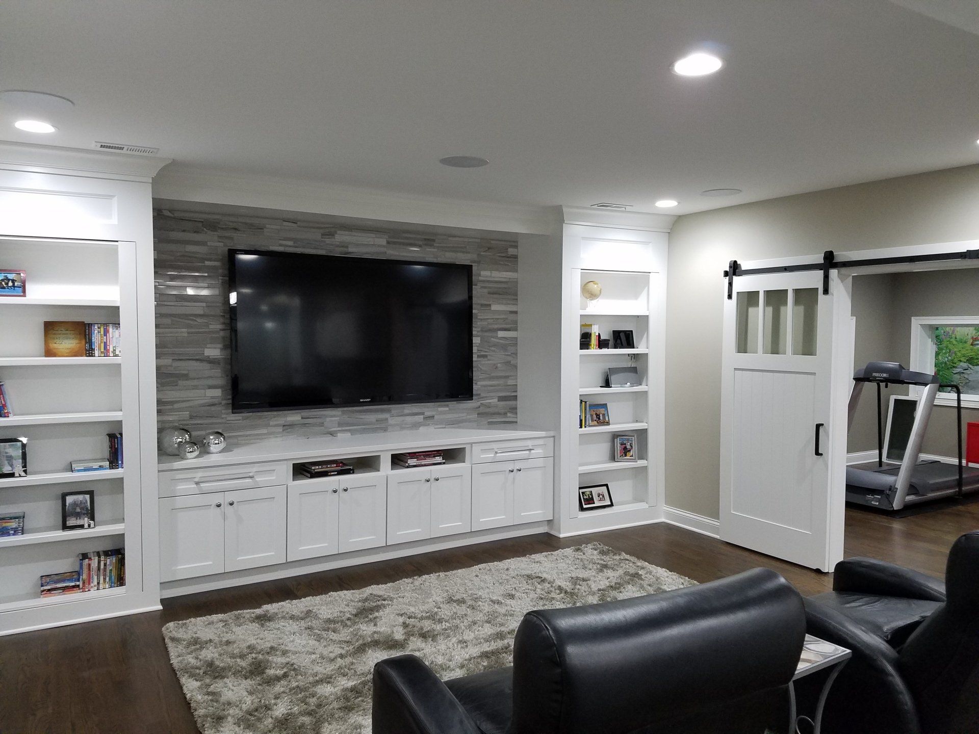 Living room with built-in white cabinets, large TV, stone accent wall, bookshelves, and a sliding barn door leading to a gym.