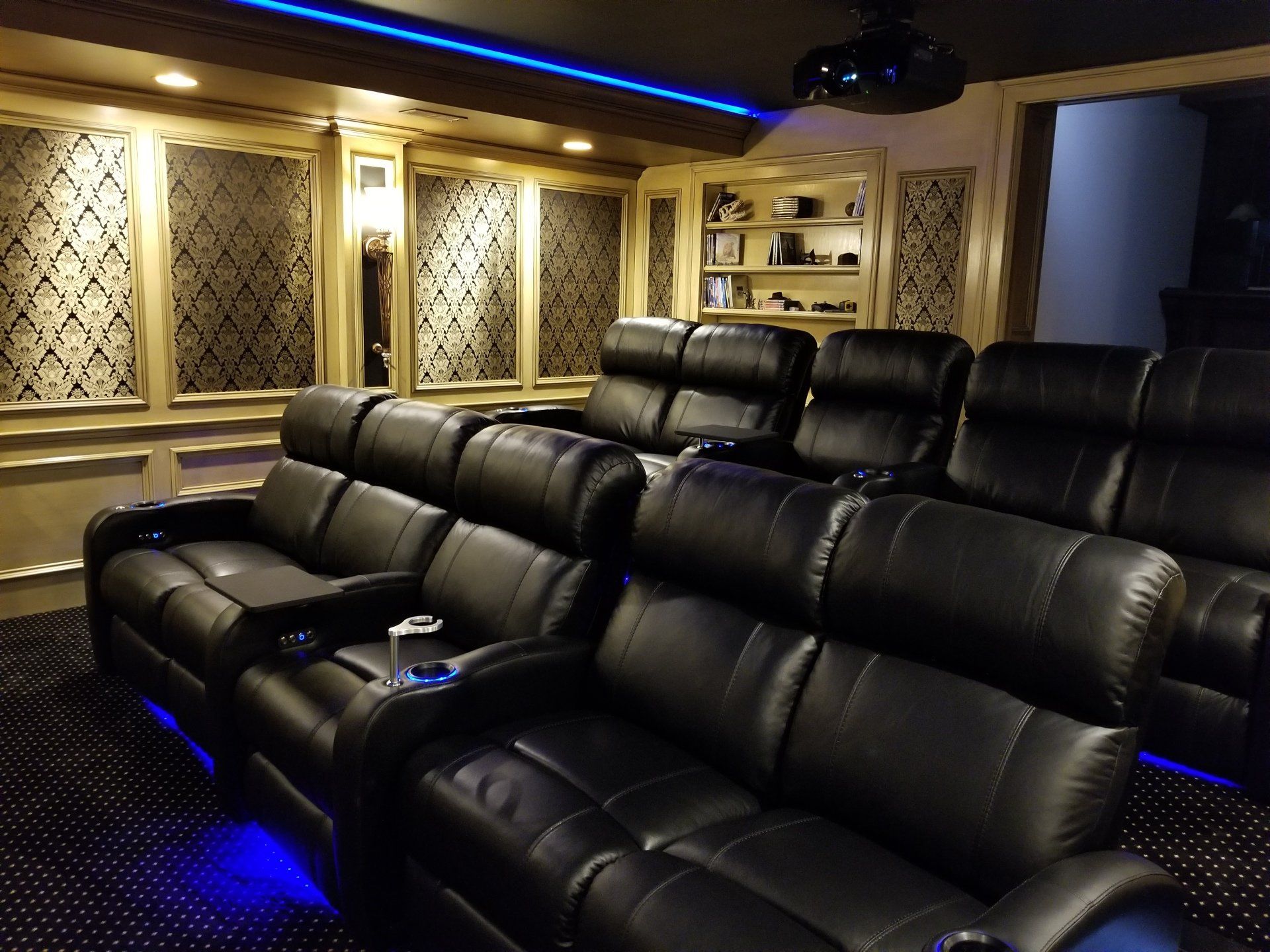 Home theater with black leather recliners, blue lighting, ornate wall panels, and a projector.