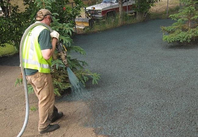 Person spraying a dark-colored surface with a hose, wearing safety gear in an outdoor setting.