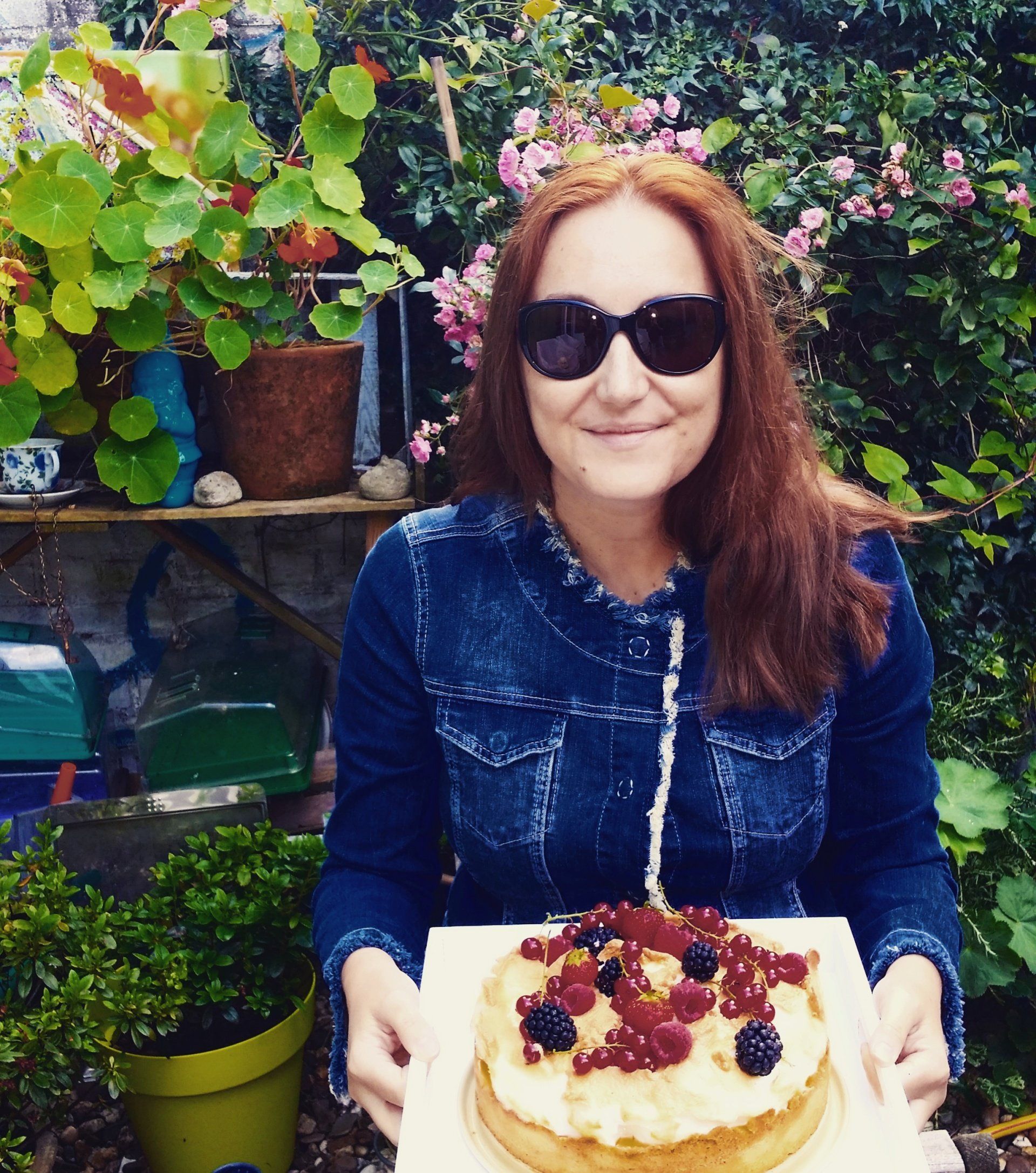 Vrouw met zonnebril die een taart met bessen vasthoudt in een weelderige tuin.