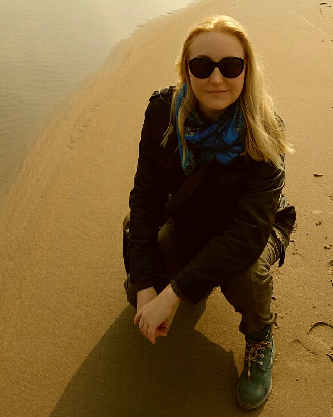 Een persoon knielt op een zandduin, draagt ​​een zonnebril, een zwarte jas, een blauwe sjaal en wandelschoenen.