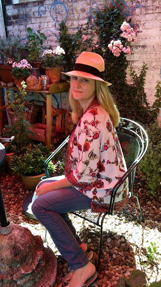 Vrouw met een strohoed zit op een smeedijzeren stoel op een bloemrijk terras in een tuin.