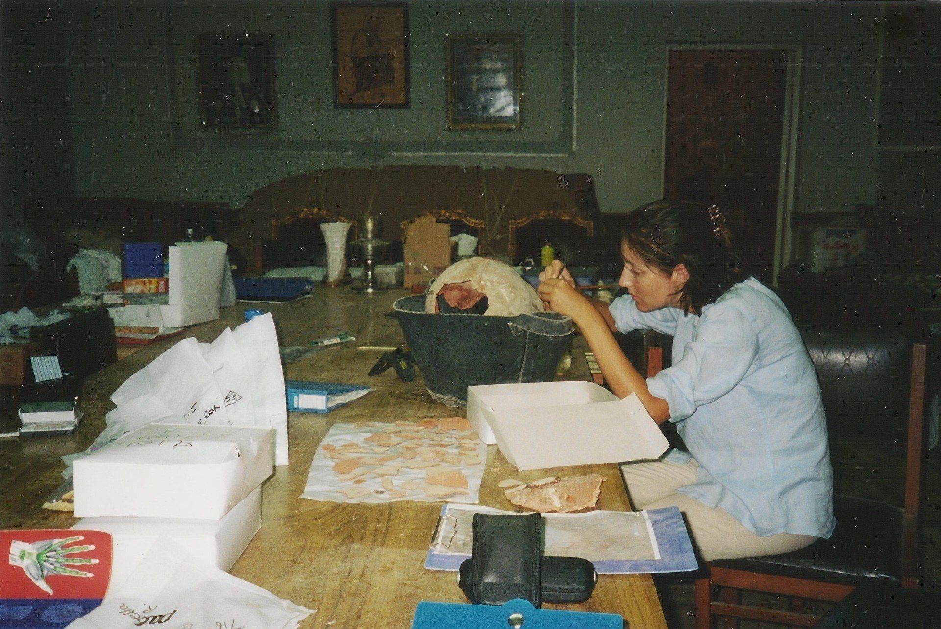 Een persoon die aan een rommelige tafel in een schemerige kamer met materialen werkt.