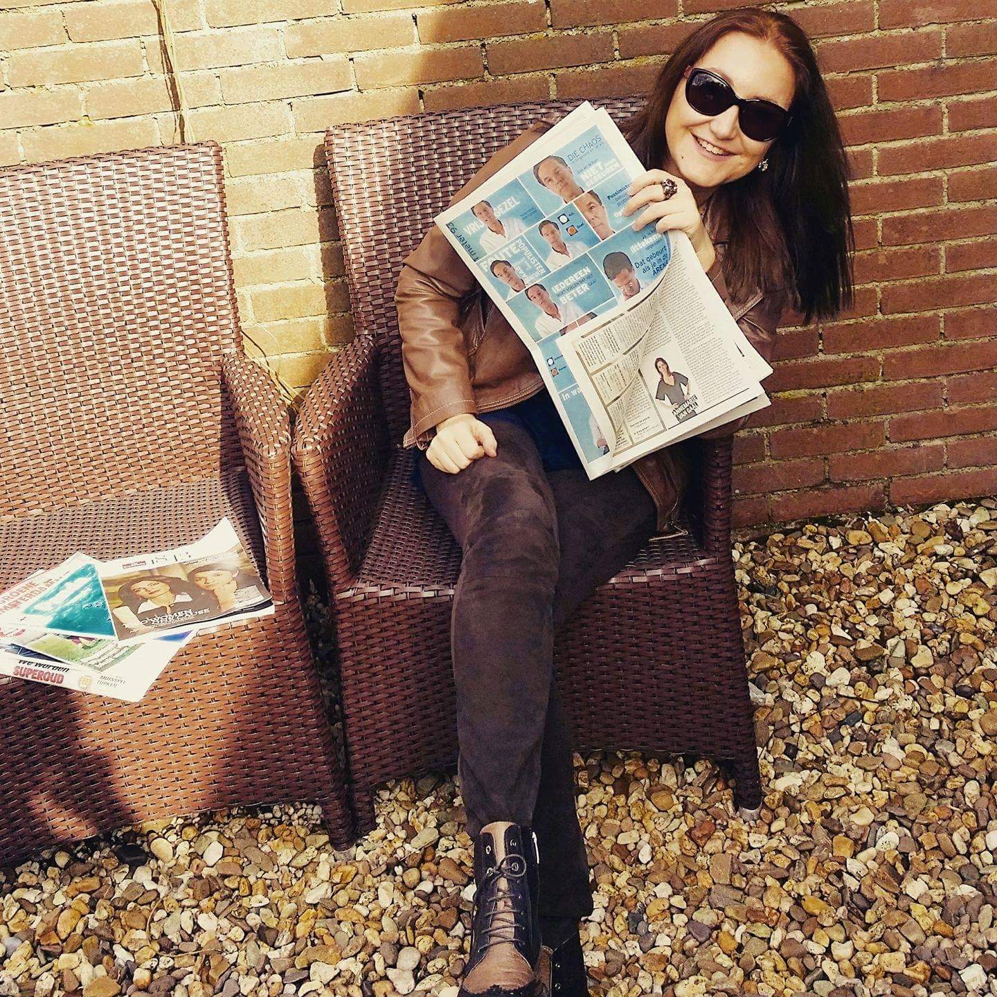 Vrouw met zonnebril leest de krant terwijl ze buiten op rieten stoelen bij een bakstenen muur zit.