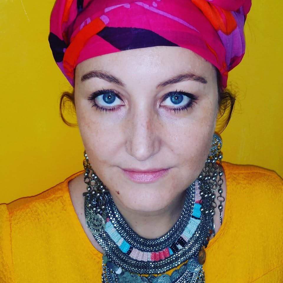 Vrouw met een felgekleurde hoofddoek en meerdere kettingen tegen een gele achtergrond, kijkt in de camera.