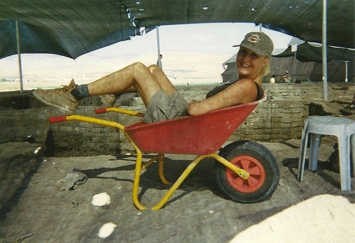 Een persoon ligt achterover in een rode kruiwagen op een zandstrand onder een afdak en lacht naar de camera.