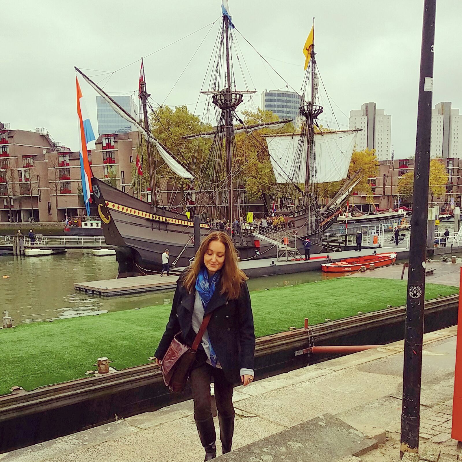Vrouw staat aan de oever van een kanaal met een historisch zeilschip en bomen op de achtergrond.