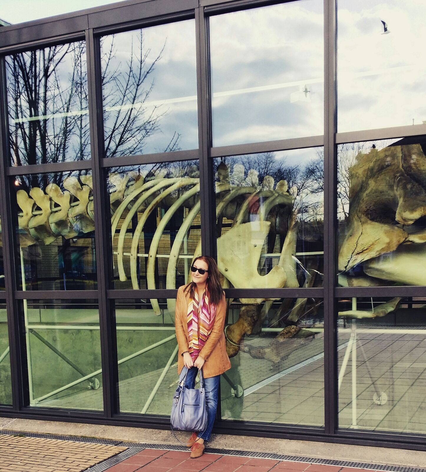 Vrouw poseert buiten een museumraam naast een tentoongesteld dinosaurusskelet.