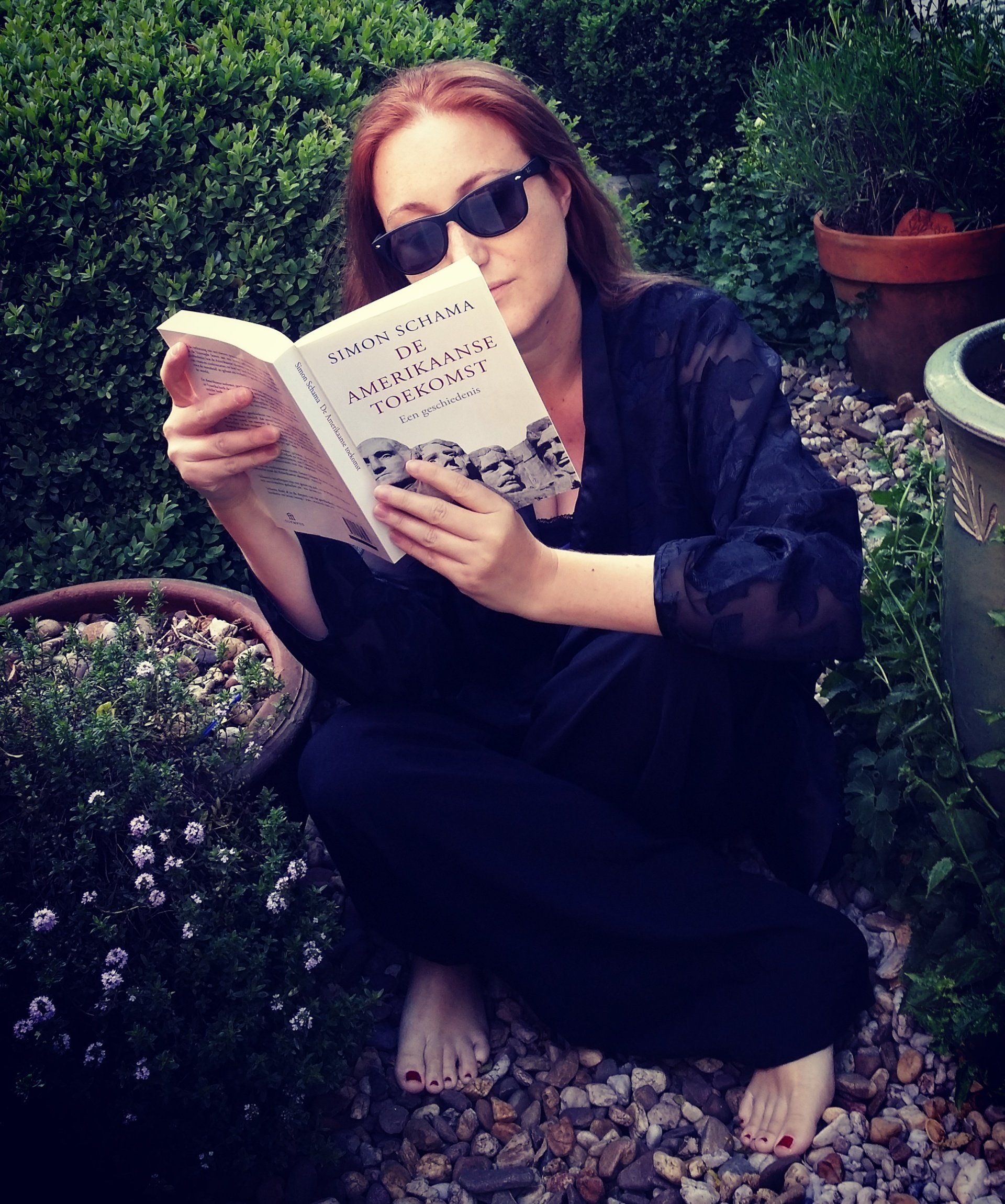 Een persoon met een zonnebril leest een boek terwijl hij in een tuin tussen potplanten zit.