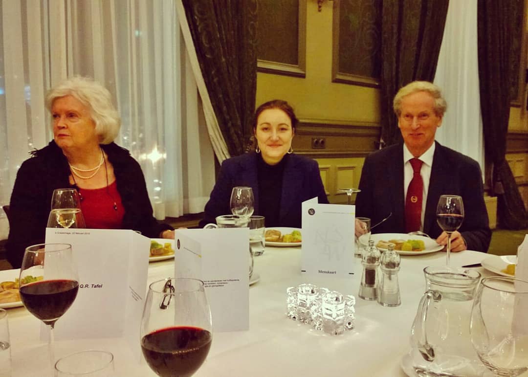 Drie personen zitten aan een formele eettafel met wijnglazen en bestek.