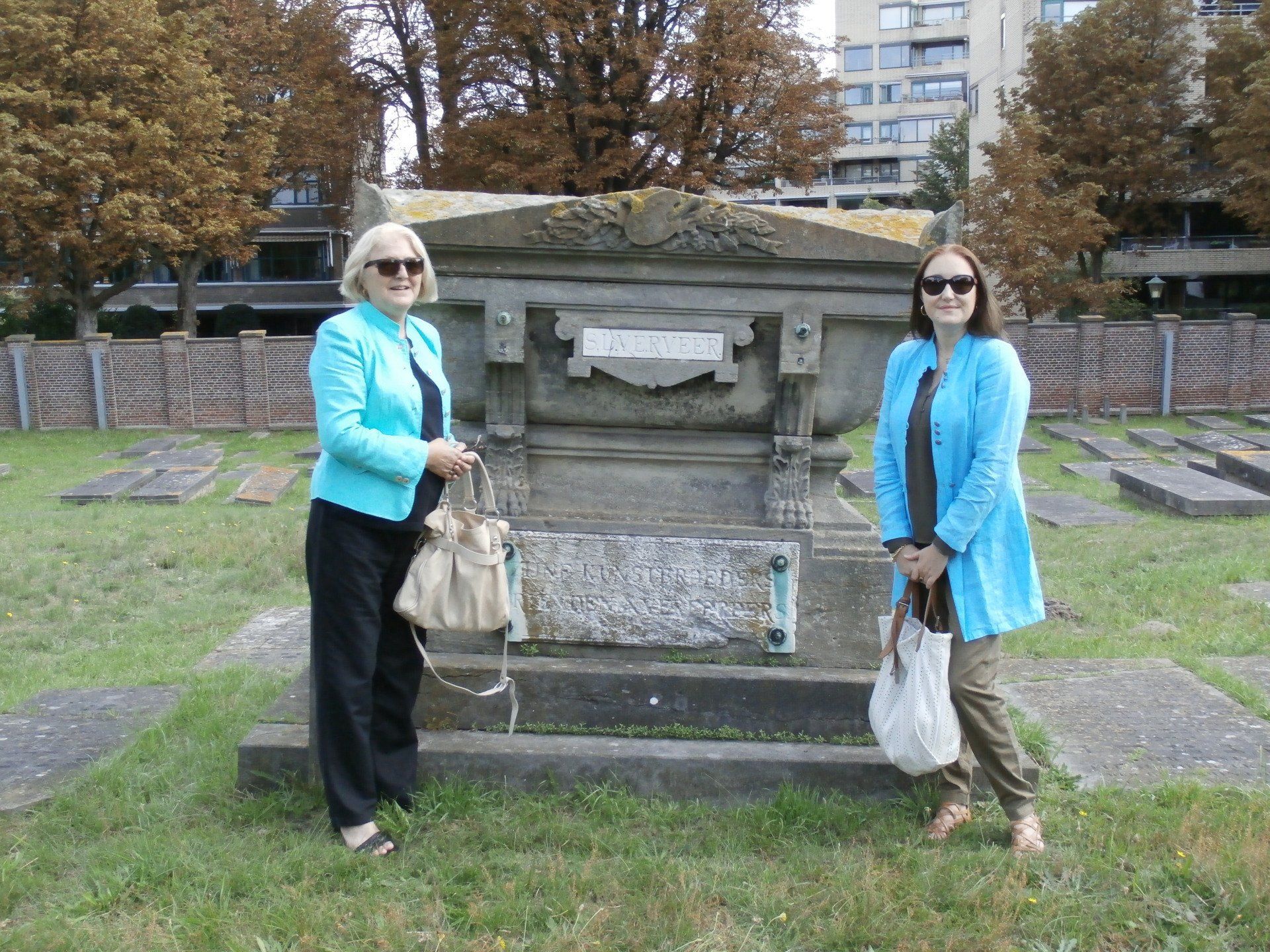 Twee mensen in blauwe jassen staan ​​naast een stenen monument in een park, met bomen en gebouwen op de achtergrond.