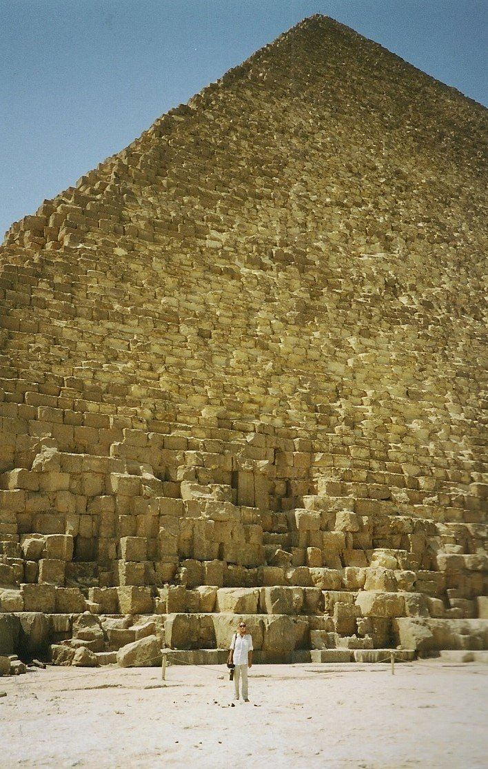 Een persoon staat voor een grote stenen piramide onder een helderblauwe hemel.