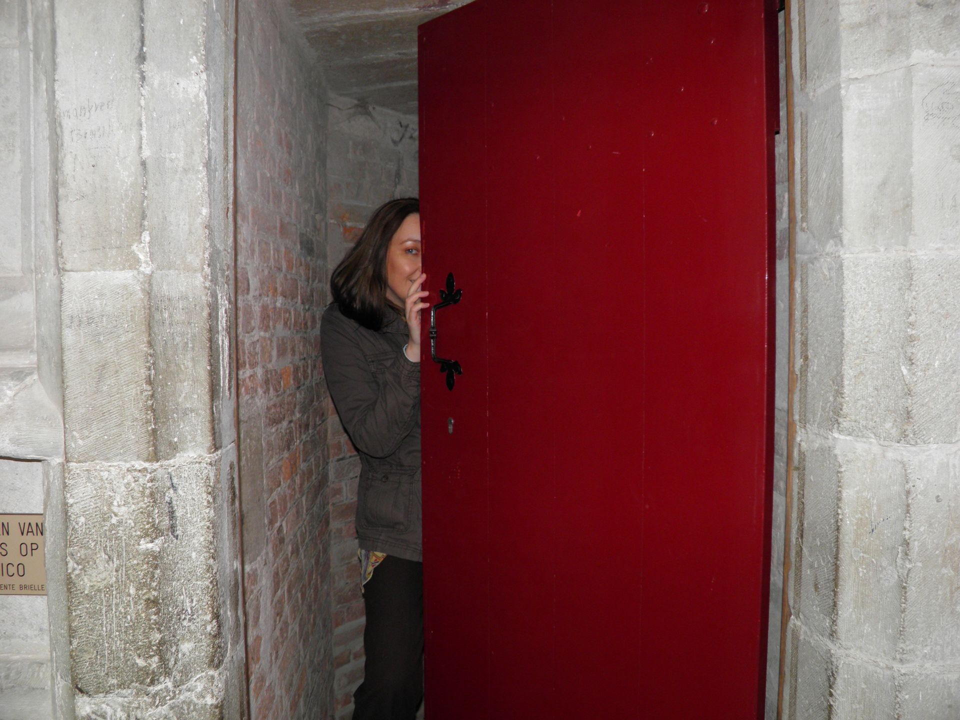 Een vrouw gluurt achter een rode deur vandaan in een grijze kamer van betonblokken.