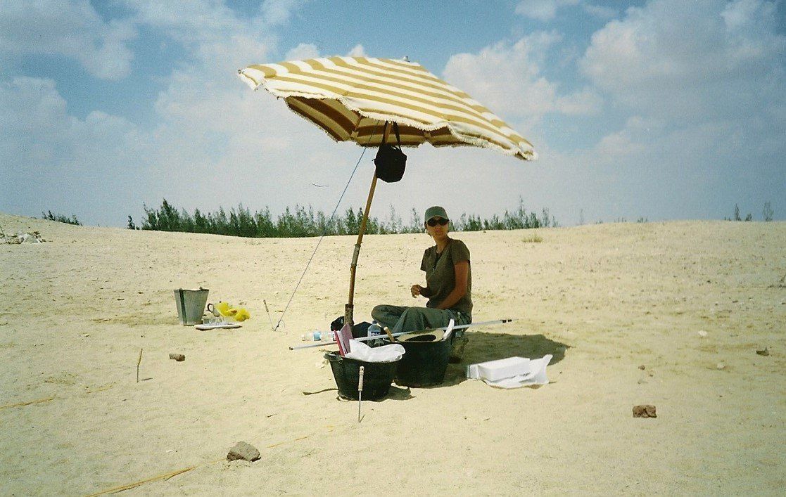 Een persoon staat onder een gestreepte parasol naast een kleine strandtent op een zandstrand.