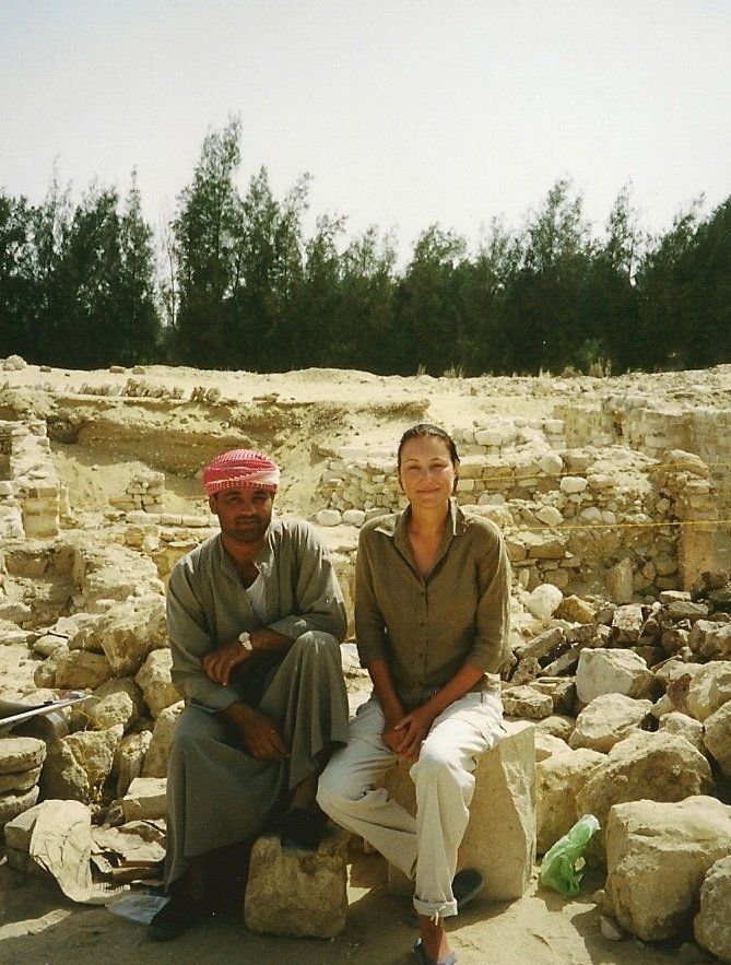 Twee mensen zitten op rotsen bij een archeologische vindplaats met stenen ruïnes en bomen op de achtergrond.