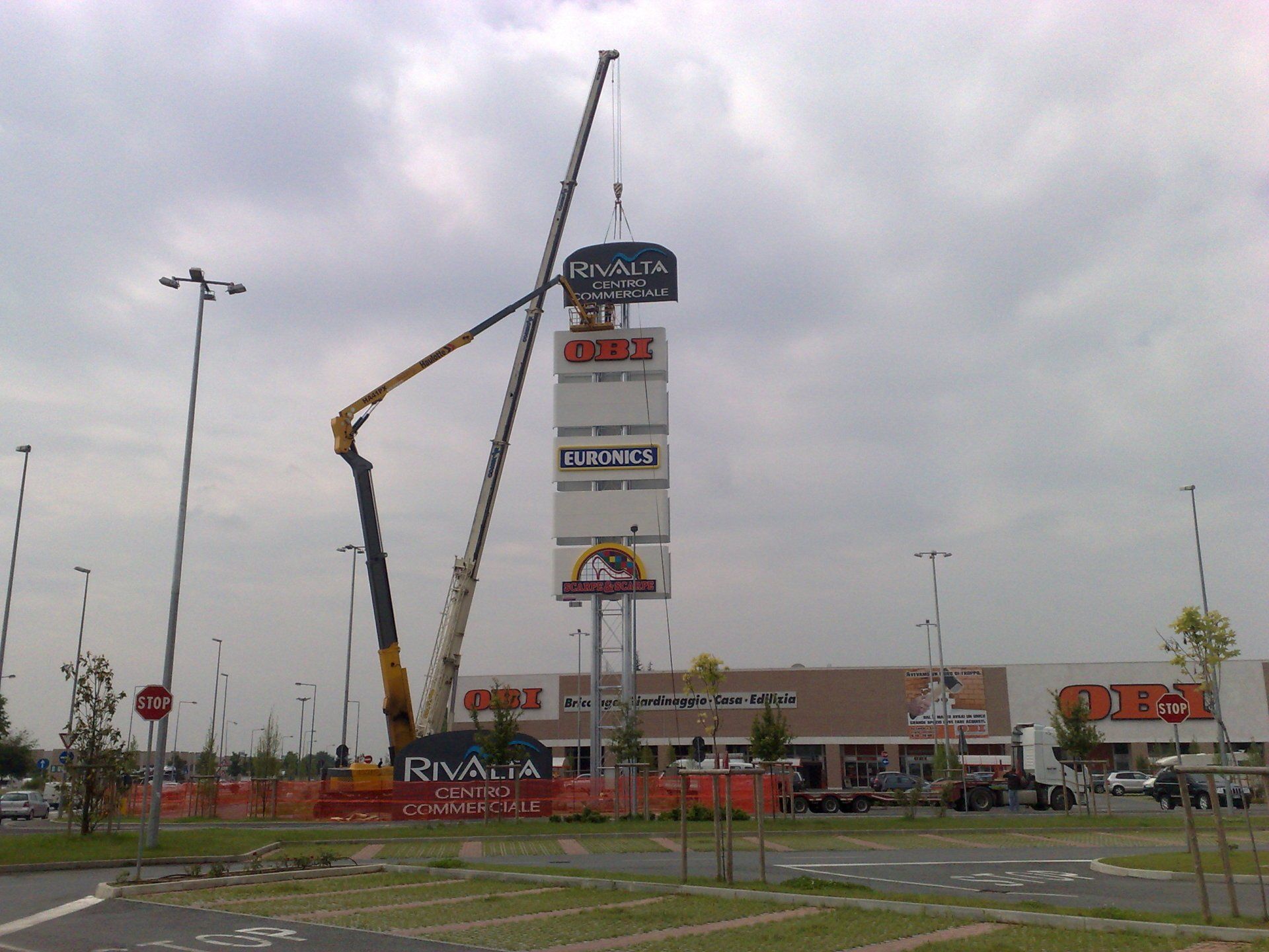 Gru che installa un grande cartello in un parcheggio. Il cartello riporta loghi e nomi.