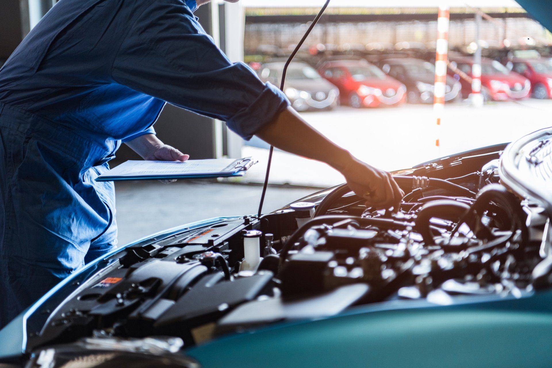 Mechanic in blue jumpsuit checks car engine in a garage, holding clipboard.