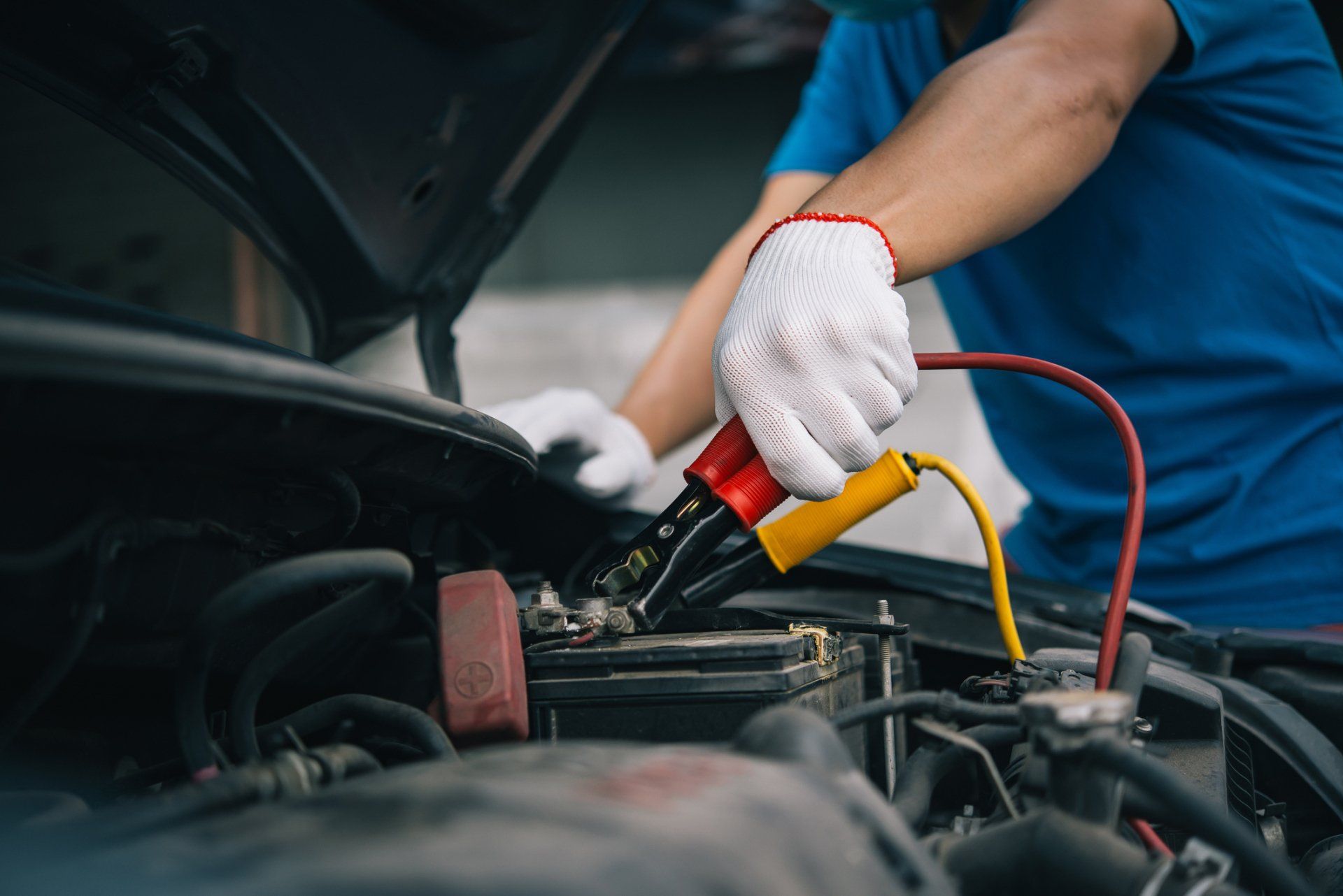 Person in gloves connecting jumper cables to a car battery under the hood.