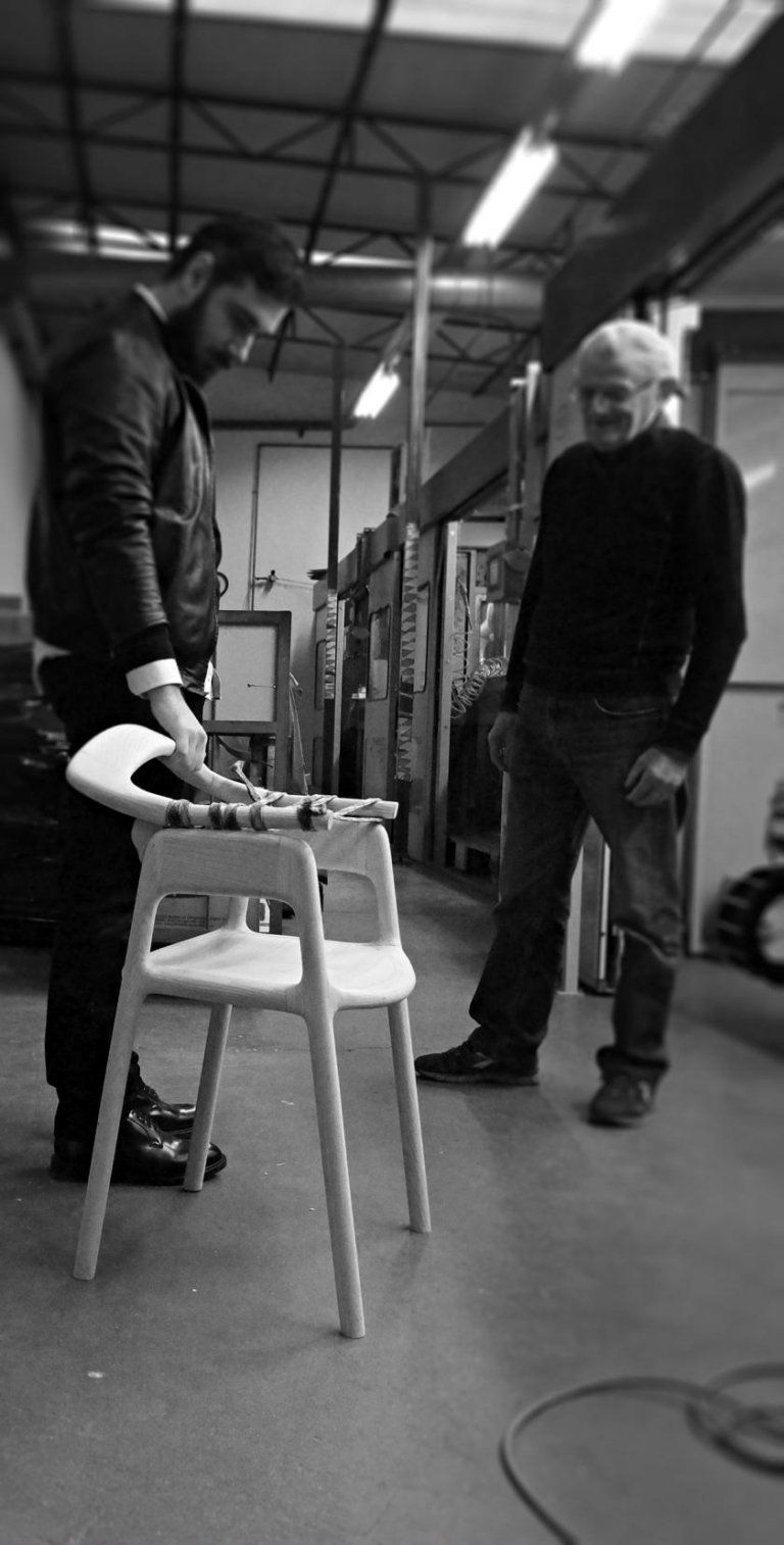Due persone in un'officina, in piedi accanto a una sedia, parlano sotto le luci del soffitto.