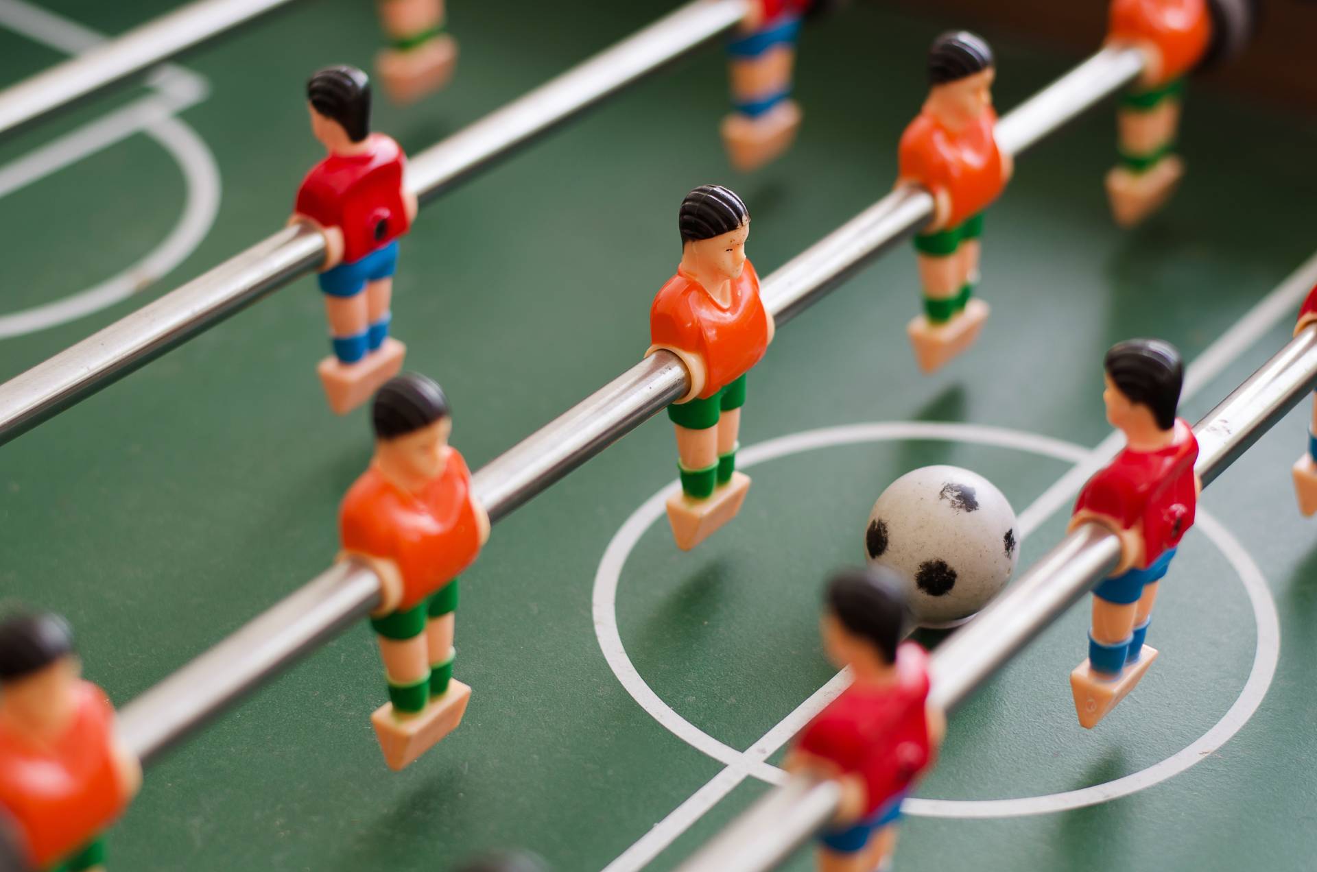 Primo piano di un biliardino con giocatori arancioni e rossi, campo verde e un piccolo pallone da calcio.